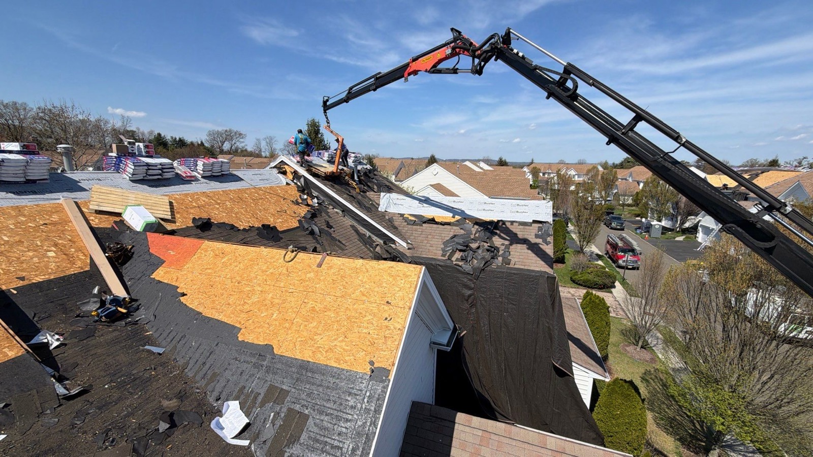 Boom crane lifting pallets of GAF shingle bundles onto a large suburban roof while LGM crew works