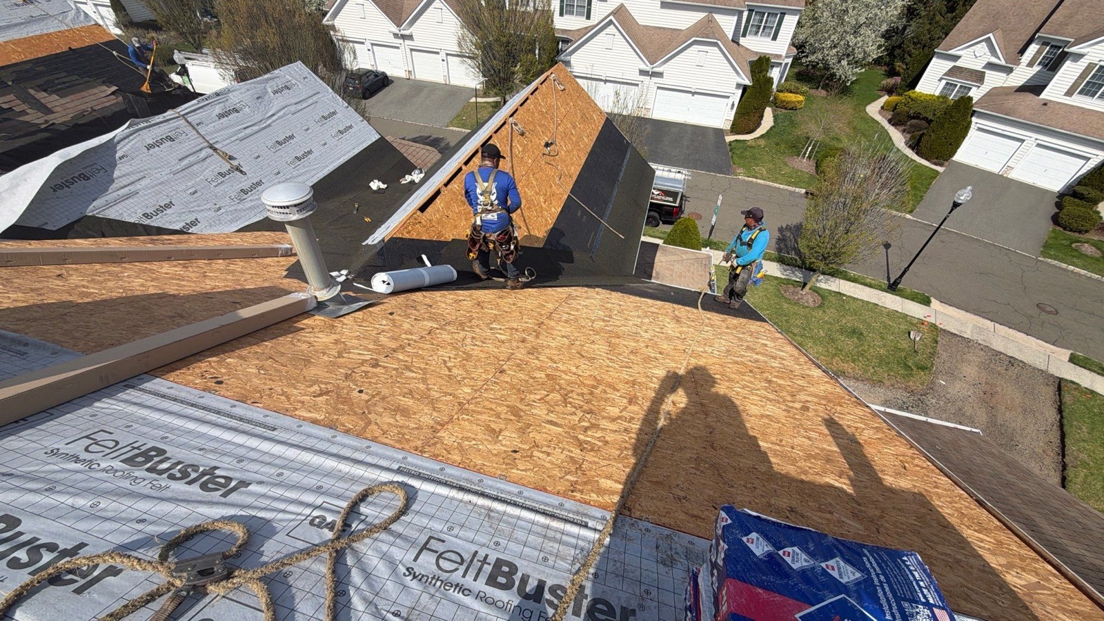 Two LGM crew members harnessed on a freshly resheathed roof deck with FeltBuster and a suburban cul-de-sac below